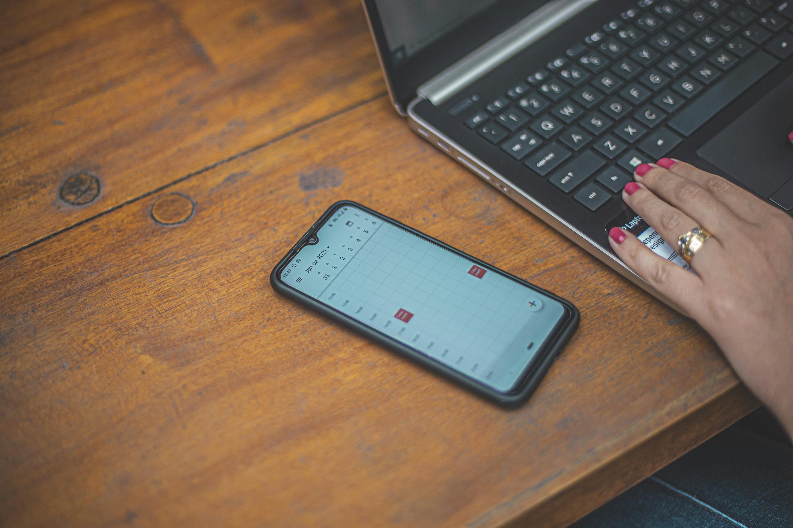 Close-up of a hand using a laptop with a smartphone showing a calendar on a wooden desk.